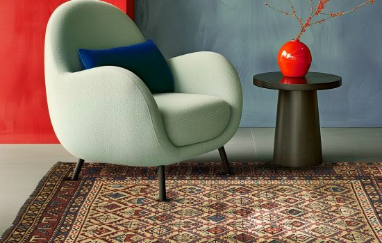 tappeto persiano antico con motivi geometrici su sfondo beige sotto poltrona di design moderno in pelle bianca, con un tavolino moderno sotto vaso rosso di design, e parete rossa alle spalle della poltroncina. Forte illuminazione di charme
